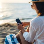 Woman using smartphone on a beach in Zakynthos to plan her holiday with AI assistance