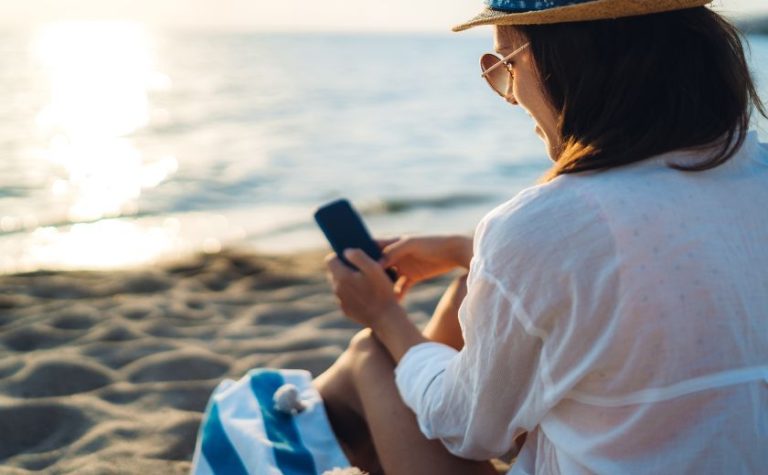 Woman using smartphone on a beach in Zakynthos to plan her holiday with AI assistance